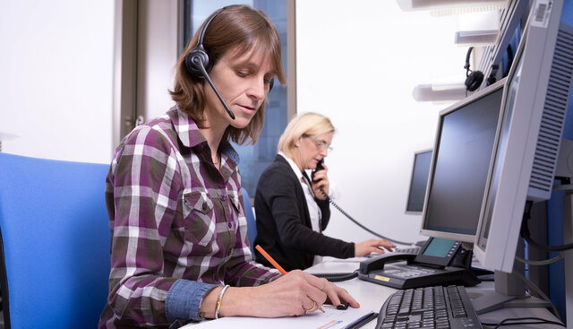 1 Ärztin und 1 Telefonistin telefonieren im Krebsinformationsdienst. Die ärztin im Vordergrund hat ein headset auf und notiert nebenher etwas; die telefonistin im Hintergrund hält den Hörer in der Hand.