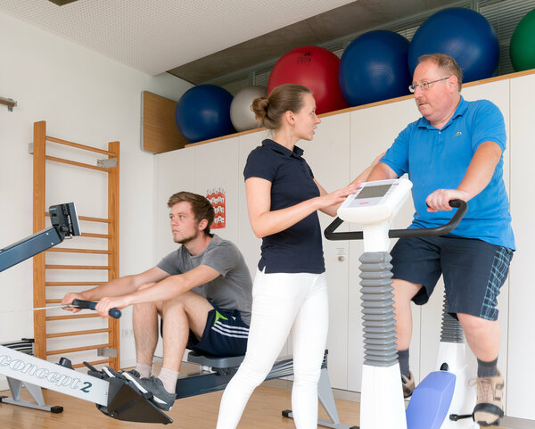 Ein Mann fährt in einem Sportraum von einer Physiotherapeutin begleitet auf dem Fahrradergometer. Im Hintergrund trainiert ein junger Mann auf einem Rudergerät.