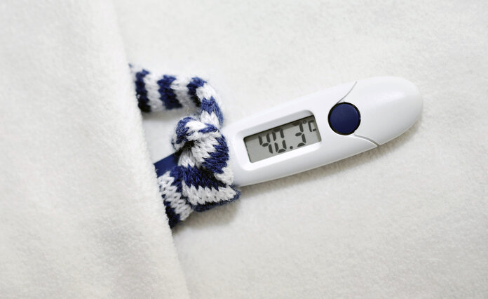 Fieberthermometer, das 40,3 Grad anzeigt, einen winzigen blau-weiß gestreiften Schal trägt und in einem winzigen weißen Bett liegt