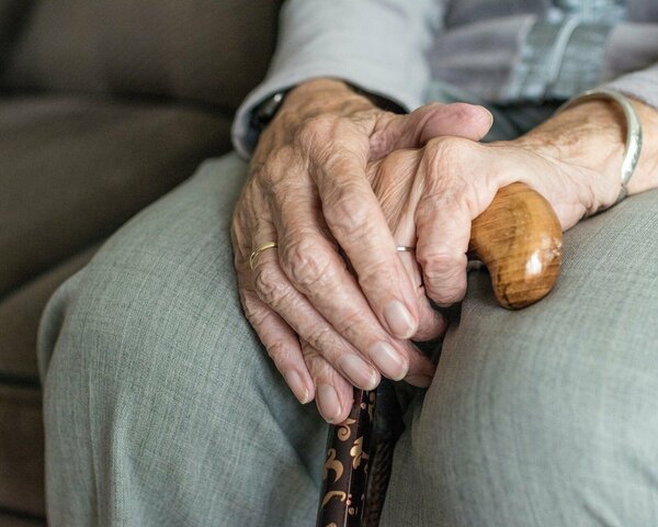Nahaufnahme einer Seniorin, die mit übereinander gelegten Händen auf dem Sofa sitzt.