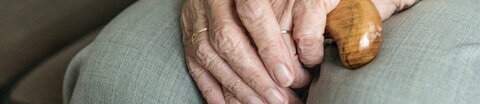 Nahaufnahme einer Seniorin, die mit übereinander gelegten Händen auf dem Sofa sitzt.