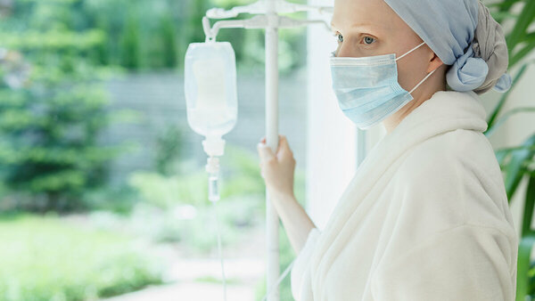 Patientin mit Kopftuch und Mundschutz hält einen Infusionsständer und schaut aus dem Fenster.