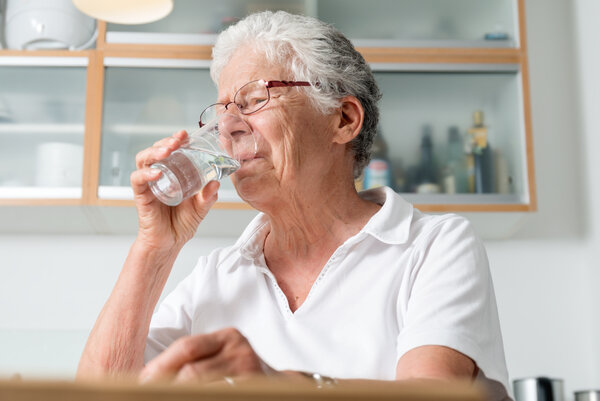 Ältere Frau sitzt in der Küche und trinkt Wasser aus einem Glas.