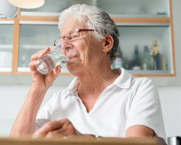 Ältere Frau sitzt in der Küche und trinkt Wasser aus einem Glas.