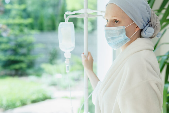 Patientin mit Kopftuch und Mundschutz hält einen Infusionsständer und schaut aus dem Fenster.