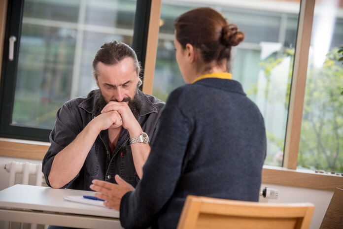 Mann sitzt bedrückt an einem Tisch, vor ihm sitzt eine Psychoonkologin