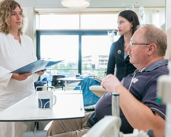 Ärztin steht vor dem Krankenbett eines Patienten mit seiner Angehörigen und spricht über die weitere Behandlung.