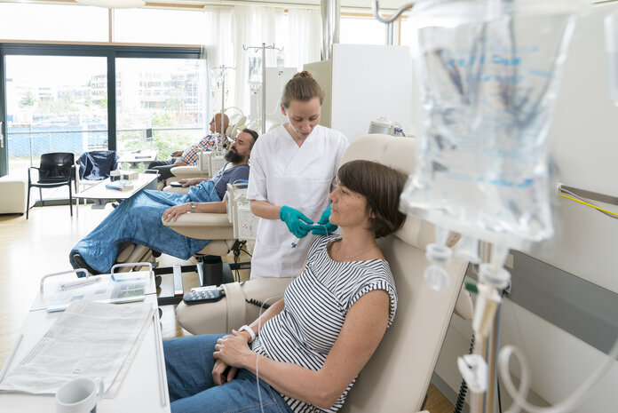 Eine Patientin sitzt in einer Chemo-Ambulanz mit anderen Patienten. Eine Krankenschwester bereitet ihre Infusion vor.