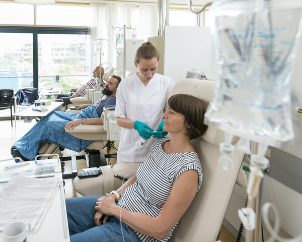 Eine Patientin sitzt in einer Chemo-Ambulanz mit anderen Patienten. Eine Krankenschwester bereitet ihre Infusion vor.