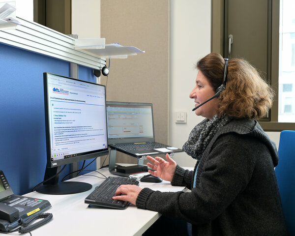 Eine Ärztin vom Telefondienst des Krebsinformationsdienstes sitzt an einem Arbeitsplatz mit 2 Computer-Bildschirmen, auf die sie schaut, während sie über ein Headset telefoniert.