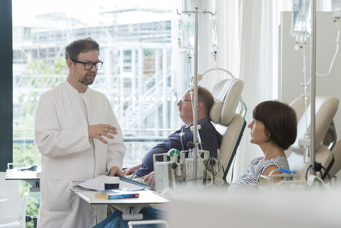 Eine Patientin und ein Patient sitzen nebeneinander in Behandlungsstühlen einer Tagesklinik und erhalten eine Infusion, ein Arzt steht bei der Patientin und erklärt etwas.