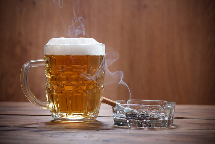 Volles Bierglas steht auf einem Tisch neben einem Aschenbecher mit einer qualmenden Zigarette